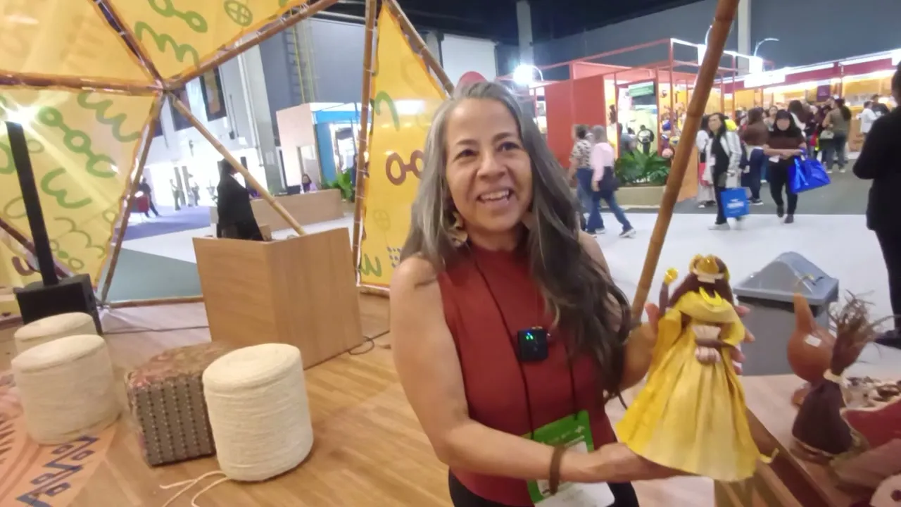 A artesã Alice de Oliveira segura uma Oxum feita de palha de milho, com vestido amarelo, no estande da feira